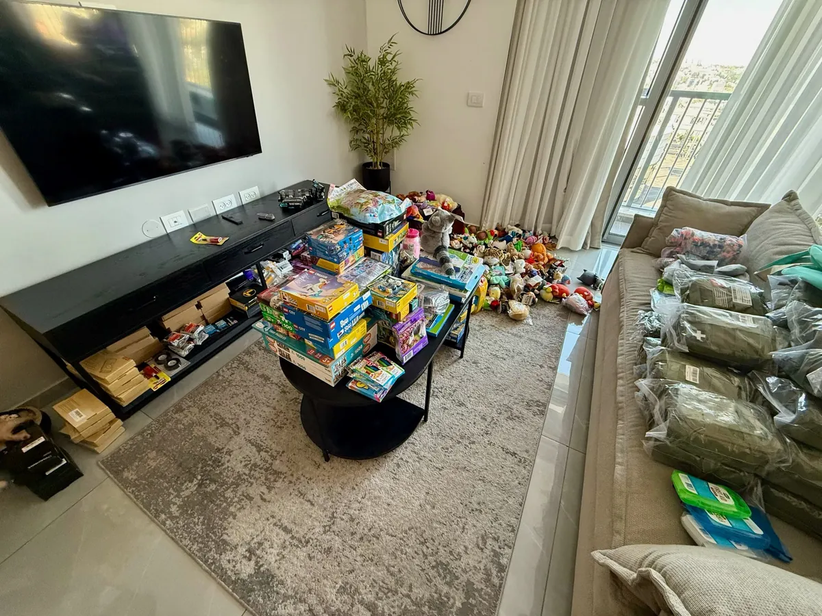 A living room filled with aid ready to travel: stacks of LEGO and board games, piles of stuffed animals, and first-aid kits in olive pouches lined up on the sofa.