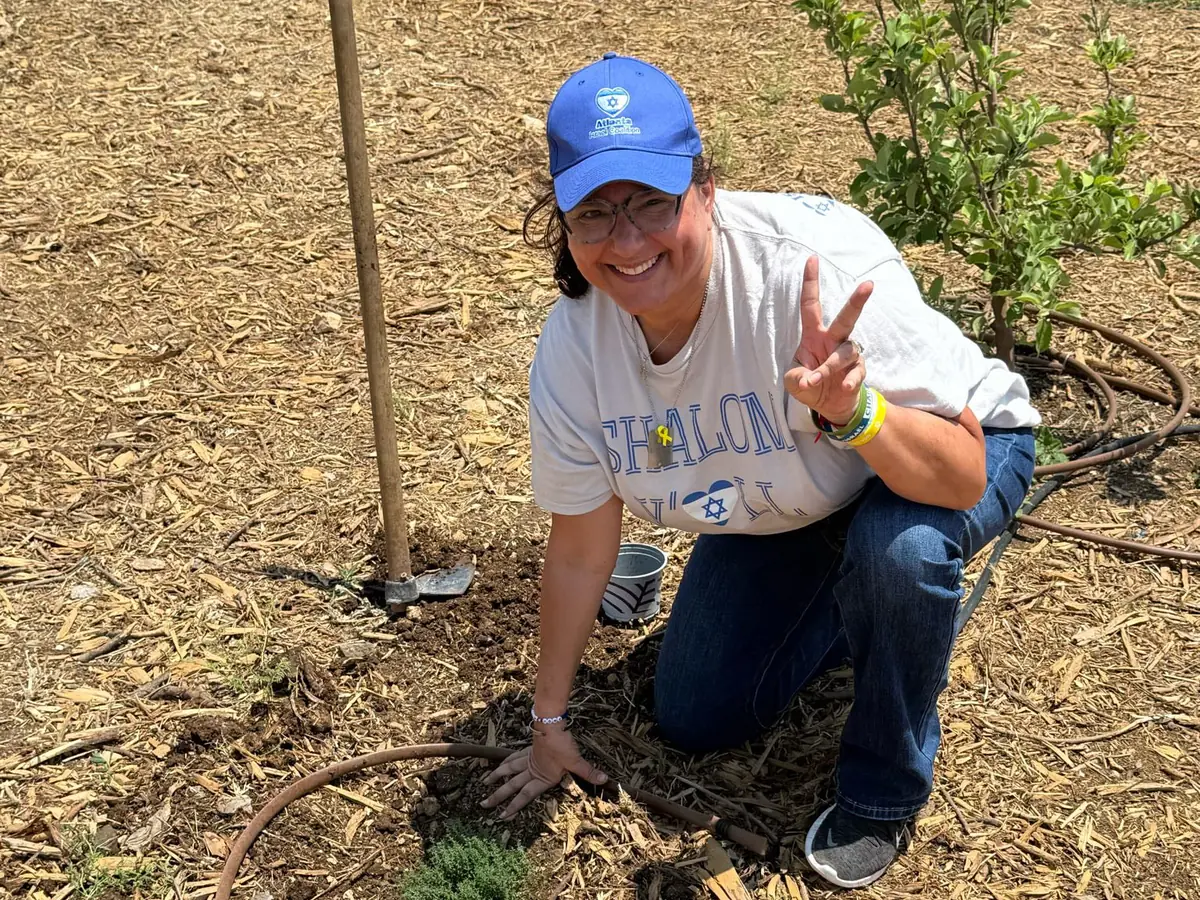 An AIC volunteer in an AIC cap kneels in the mulch beside a newly planted seedling and gives a peace sign.