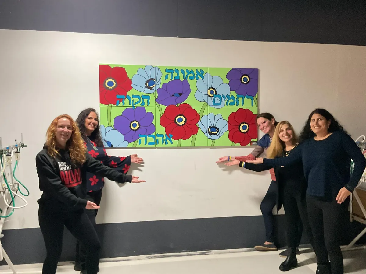 Five AIC volunteers point to a painted mural of red, blue, and purple anemones with Hebrew words for faith, hope, love, and compassion, hung on a hospital wall.