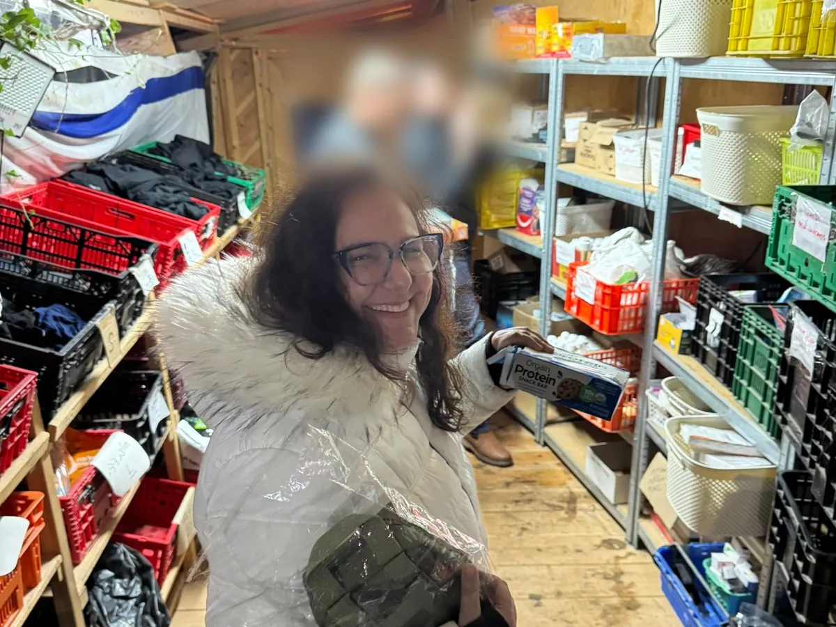 Cheryl Dorchinsky smiles in the Achim Shuva supply closet, holding a box of protein bars, shelves of crates behind her.