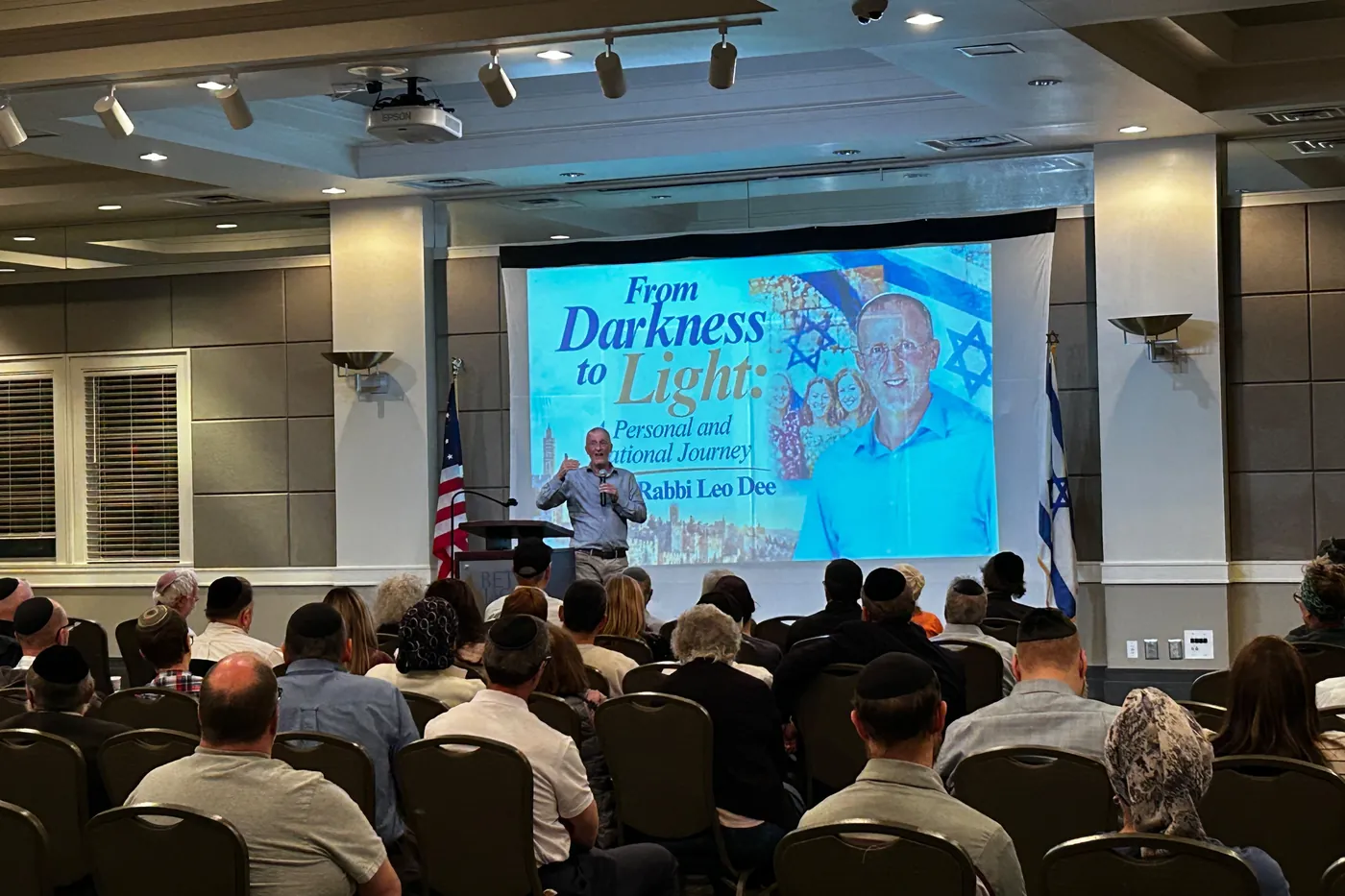 Rabbi Leo Dee speaking into a handheld microphone in front of a large screen showing the evening's title, From Darkness to Light, with an American flag and an Israeli flag on either side of the stage and the audience seated in rows in the foreground, seen from behind.