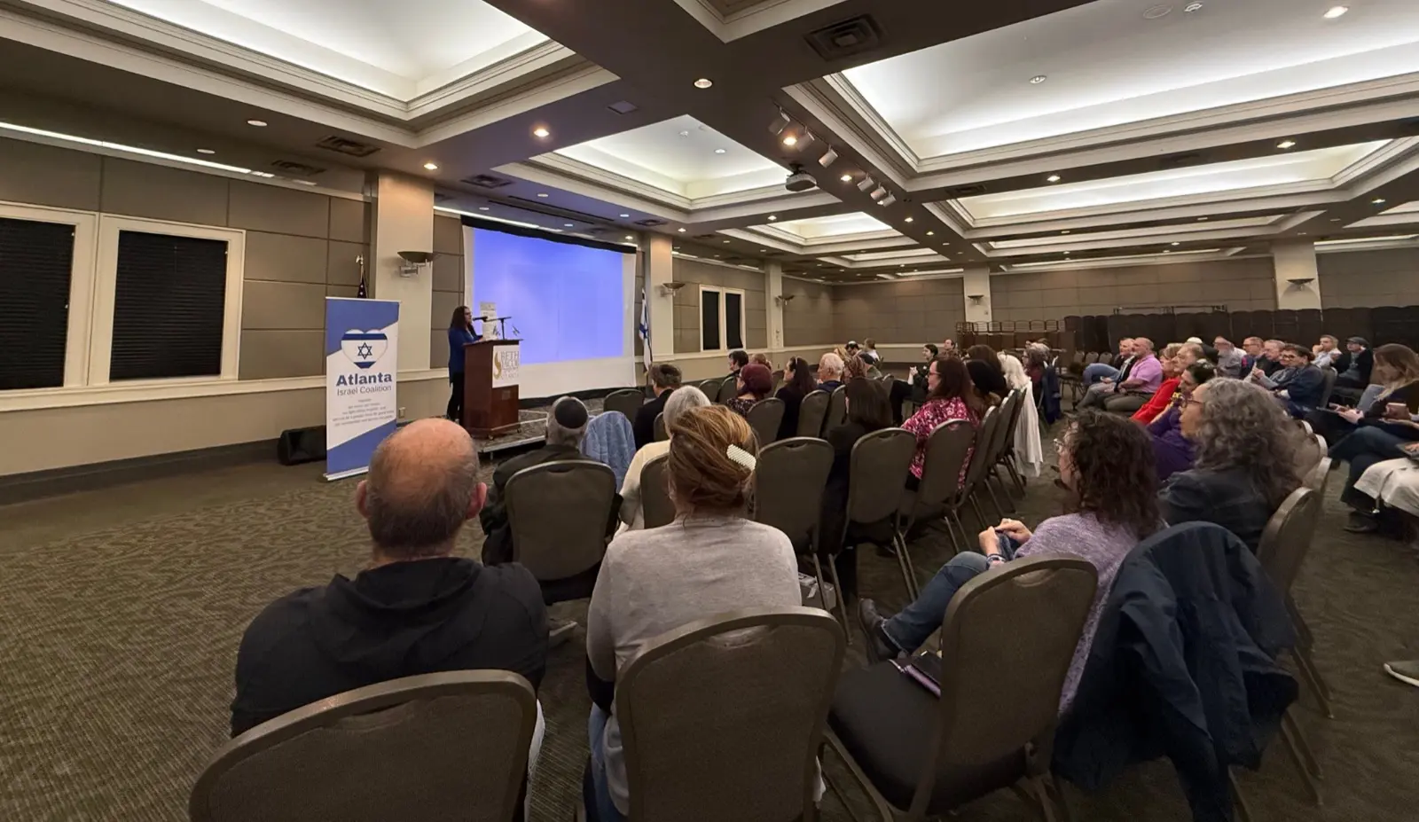 An Atlanta Israel Coalition community briefing in progress — a seated audience facing a speaker at the podium beside the AIC banner.