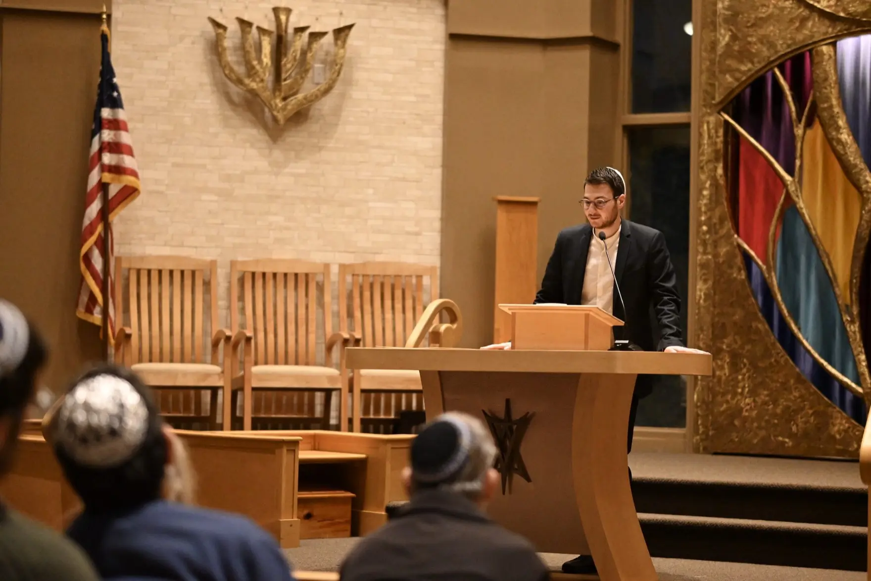 A speaker addresses a seated audience from the lectern at a synagogue, an American flag and a menorah sculpture behind them.